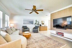 Una sala de estar moderna con ventilador de techo, televisor de pantalla plana, sofá, dos sillones, una mesa de centro y un tapete estampado.