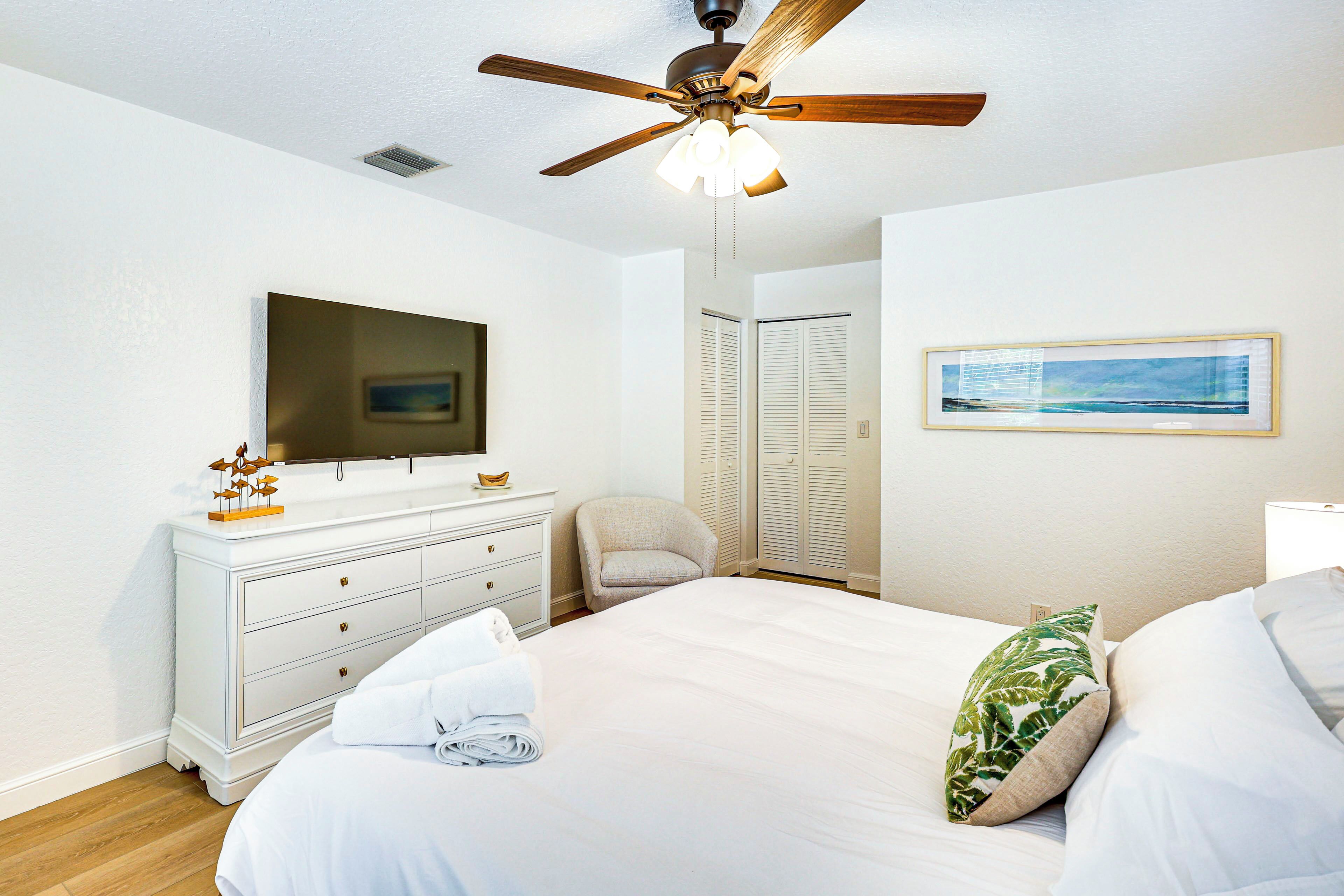 A bedroom with a bed, a dresser, a television, a ceiling fan, a chair, a closet, and a framed picture on the wall.