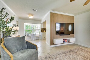 A living room with a grey sofa, a flat-screen TV on a wooden wall, a dining area with a table and chairs, and a kitchen in the background.