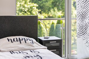 A bedroom with a bed, a bedside table, a lamp, and a window with a view of greenery.