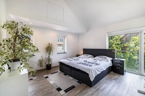 A modern bedroom with a large bed, bedside tables, a potted plant, and a view of greenery through a large window.