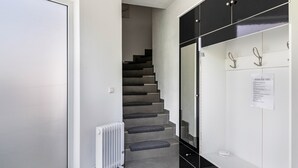 Un couloir avec une porte blanche, un escalier dont les marches sont recouvertes d’une moquette grise, un radiateur blanc et un meuble noir muni de crochets.