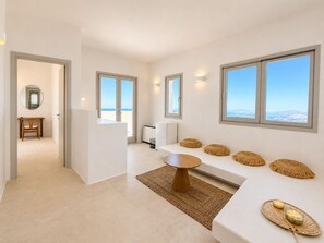 A modern interior with a sofa, a small table, and a view of the sea through large windows. - Vespera Private Luxury Suites (Santorini)