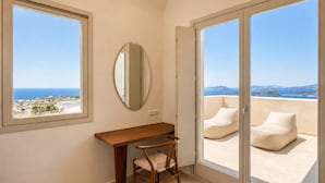 A room with a desk, chair, and a large window with a view of the sea and distant land. - Vespera Private Luxury Suites (Santorini)