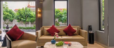 A modern living room with a beige sofa, red cushions, a coffee table with a vase and fruit, and large windows with a view of greenery.