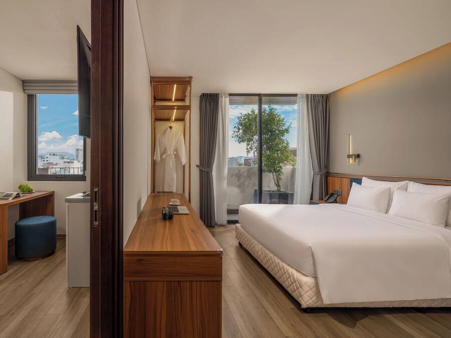A hotel room with a bed, a desk, a chair, a small table, a window with a view of buildings, and a door.