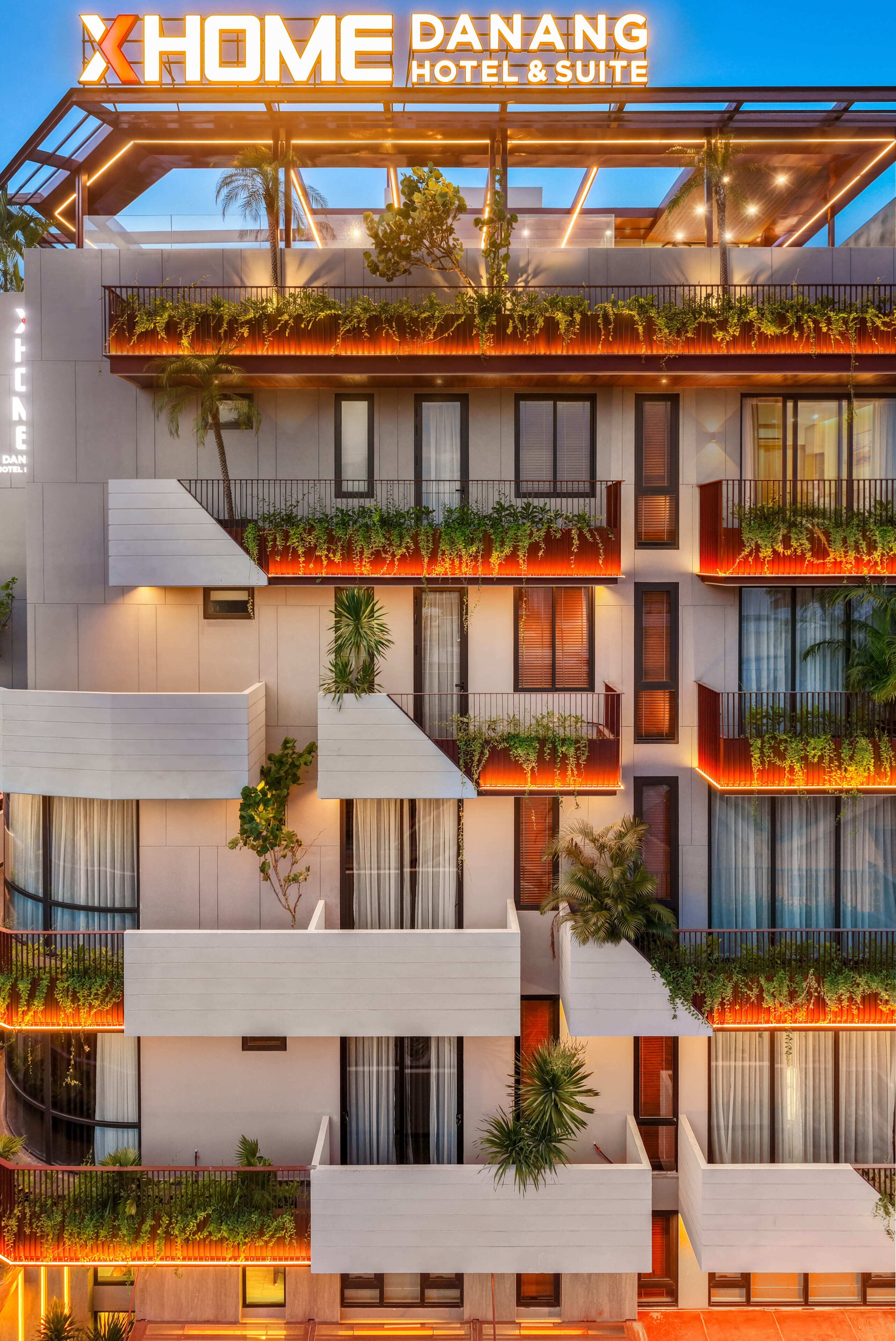 A modern hotel with multiple balconies, each adorned with plants and illuminated by warm lighting.
