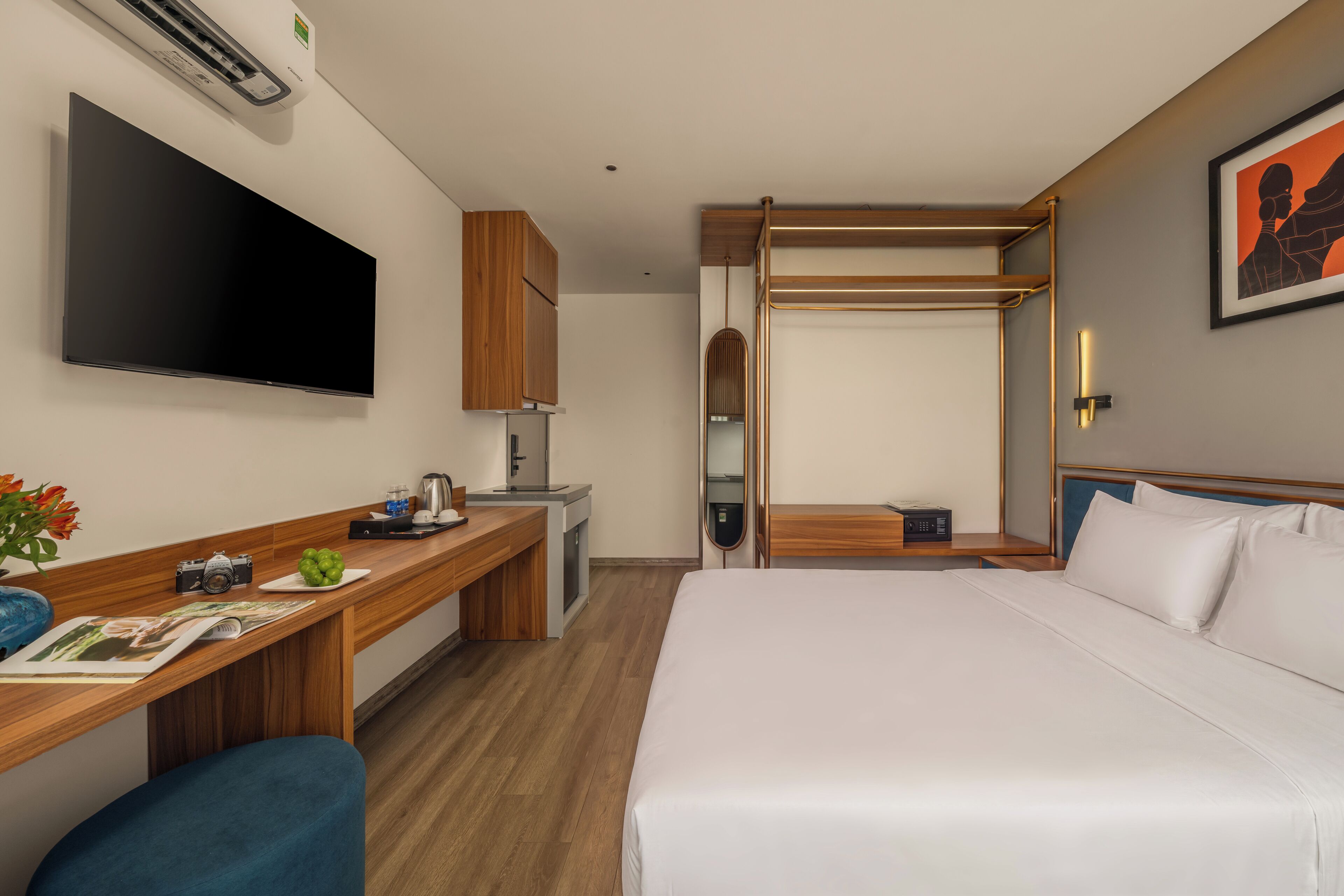 A hotel room with a bed, a desk with a flat-screen TV, a wooden desk, a chair, a small table with a kettle, a coffee machine, a mirror, and a framed picture on the wall.