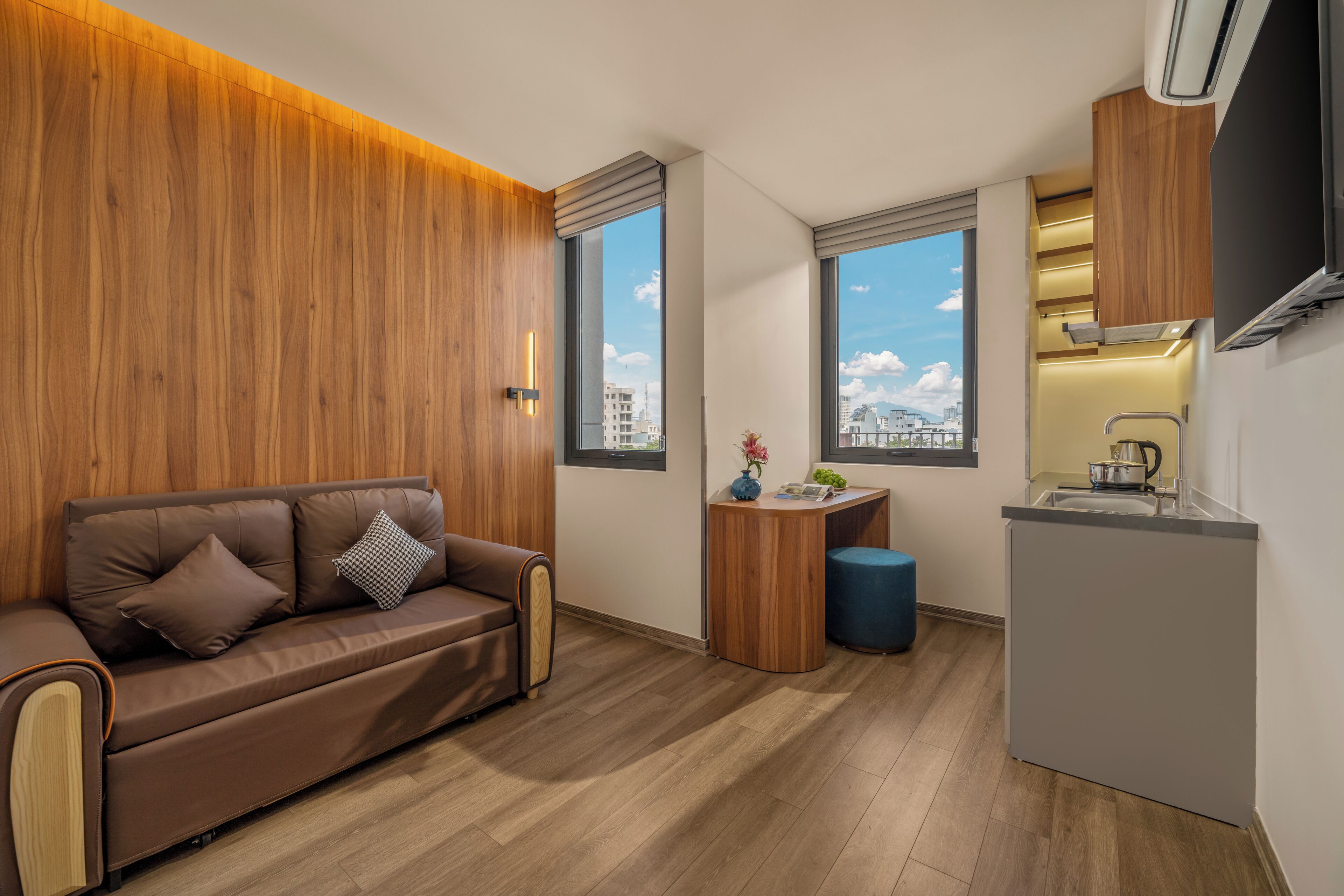 A compact living space with a sofa, a small wooden side table, a kitchenette, and a view of the cityscape through the windows.
