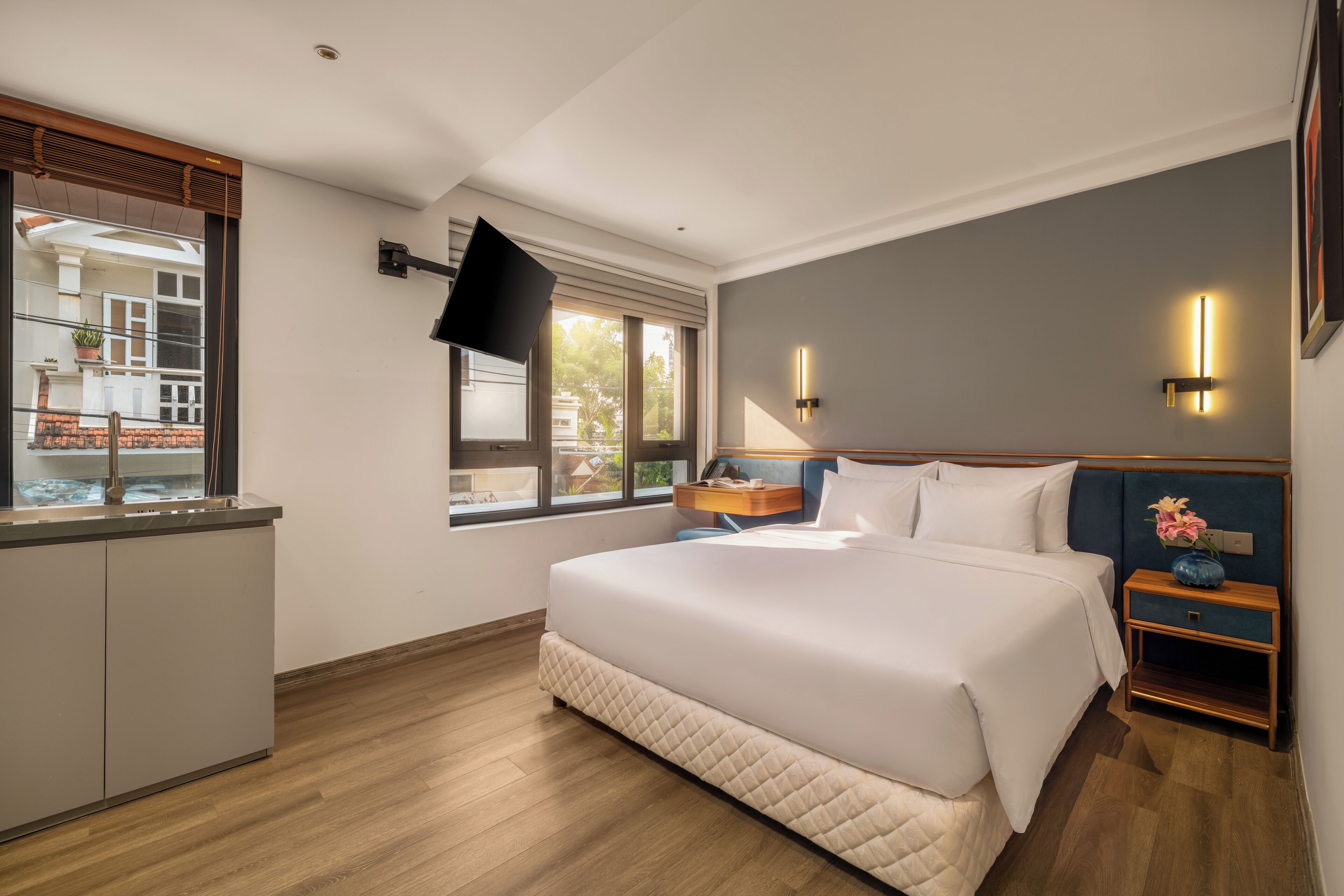 Habitación de hotel con cama, mesita de noche, televisor montado en la pared y ventana con vista a edificios.