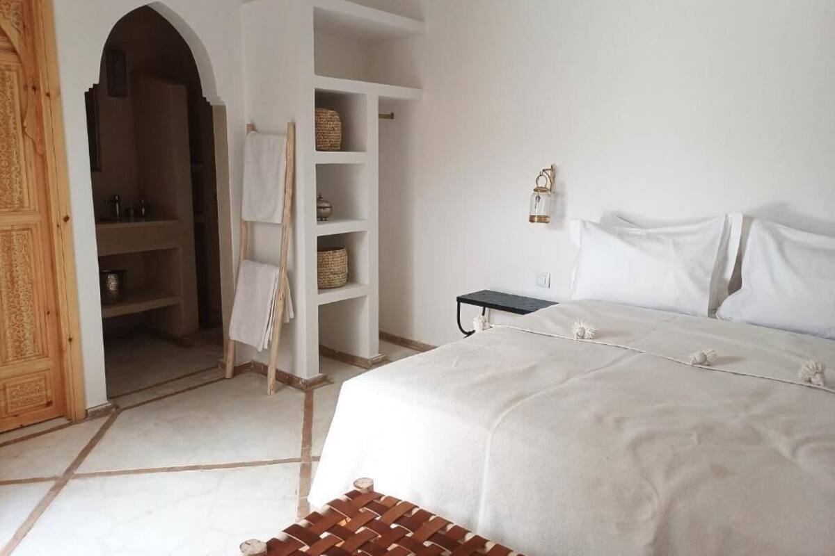 A bedroom with a bed, a wardrobe, a shelf with baskets, and a small table with a lamp.