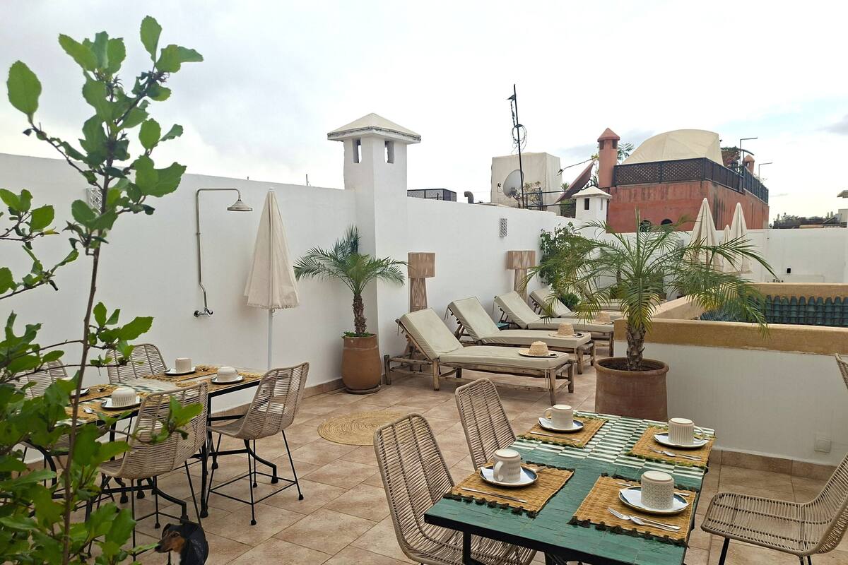 A rooftop terrace with a dining table set for four, lounge chairs, potted plants, and a white wall with a tower-like structure.