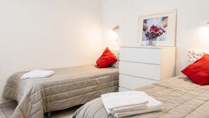 A hotel room with two beds, each with a beige quilted bedspread and red pillows. There is a white bedside table with a framed picture above it.