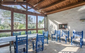 Ein überdachter Außenbereich mit Tisch und blauen Stühlen, großen Fenstern und Blick ins Grüne.