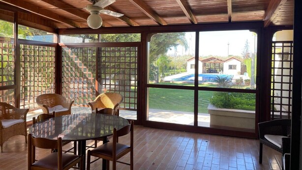 Una stanza con soffitto in legno, una ventola a soffitto, un tavolo di vetro con sedie e ampie finestre con vista su una piscina e una casa.