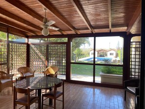 Une pièce avec un plafond en bois, un ventilateur de plafond, une table en verre avec des chaises, et de grandes fenêtres donnant sur une piscine et une maison.
