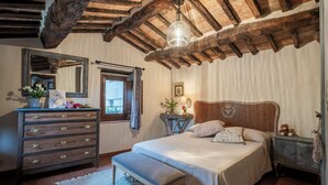 Un dormitorio con techo de madera, una cama con cabecera, un aparador, un espejo, una ventana con cortinas y una mesita de noche con lámpara.