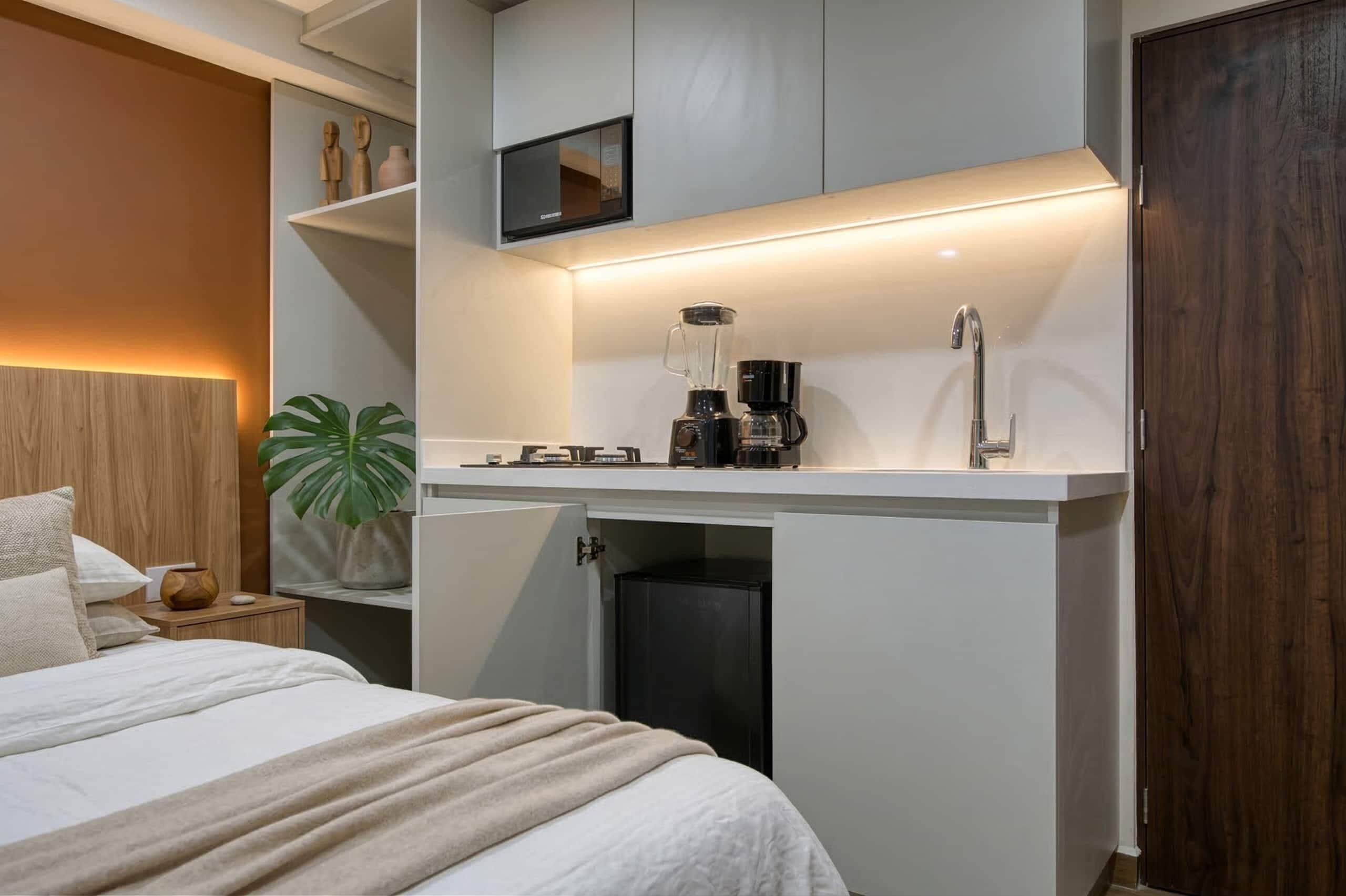 Una habitación de hotel compacta con una cama, una kitchenette pequeña, una planta y una puerta de madera.