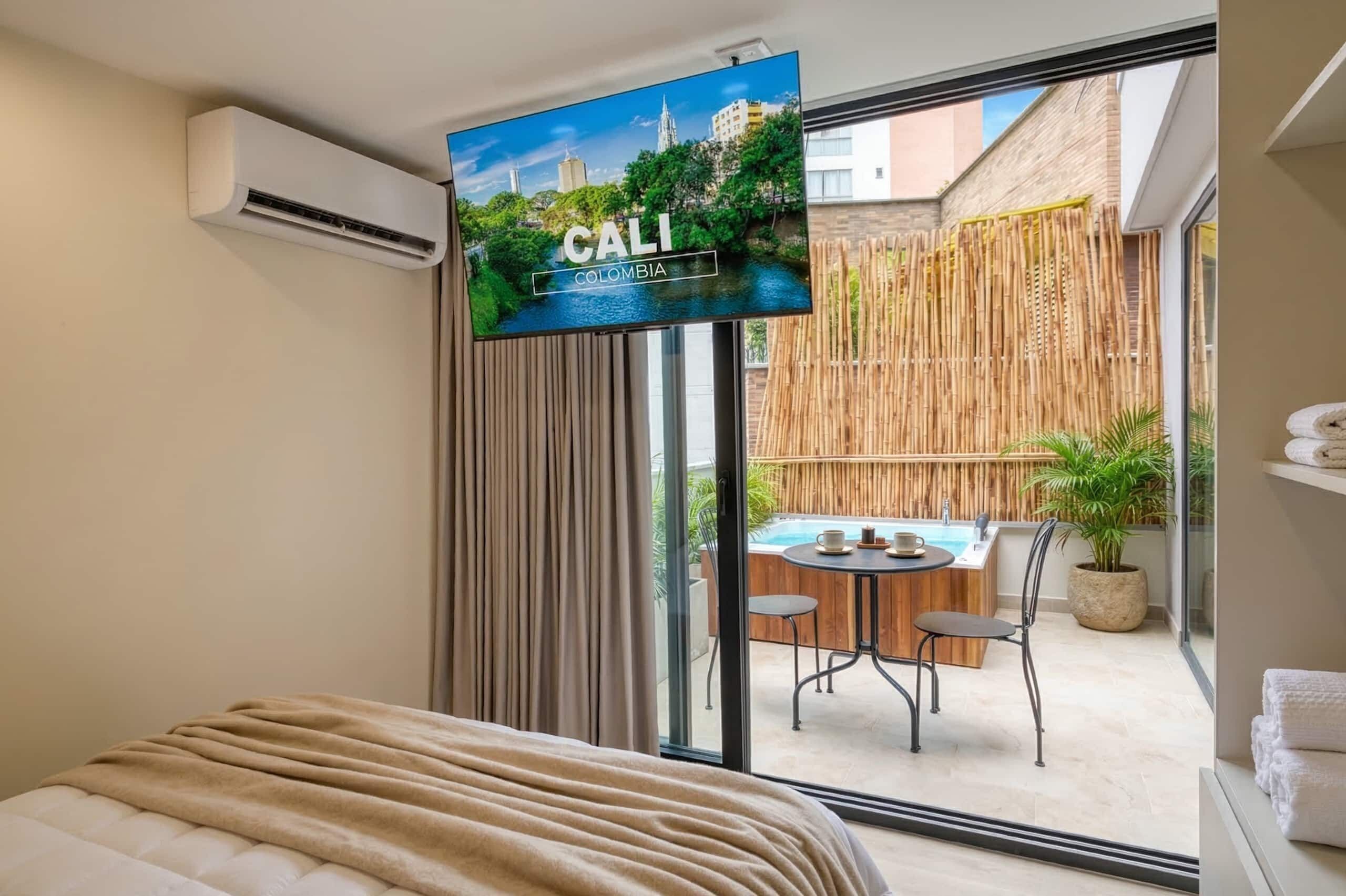 A hotel room with a bed, a television displaying a scenic view of Colombia, a bamboo fence, and a small outdoor seating area with a table and chairs.