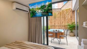 A hotel room with a bed, a television displaying a scenic view of Colombia, a bamboo fence, and a small outdoor seating area with a table and chairs.