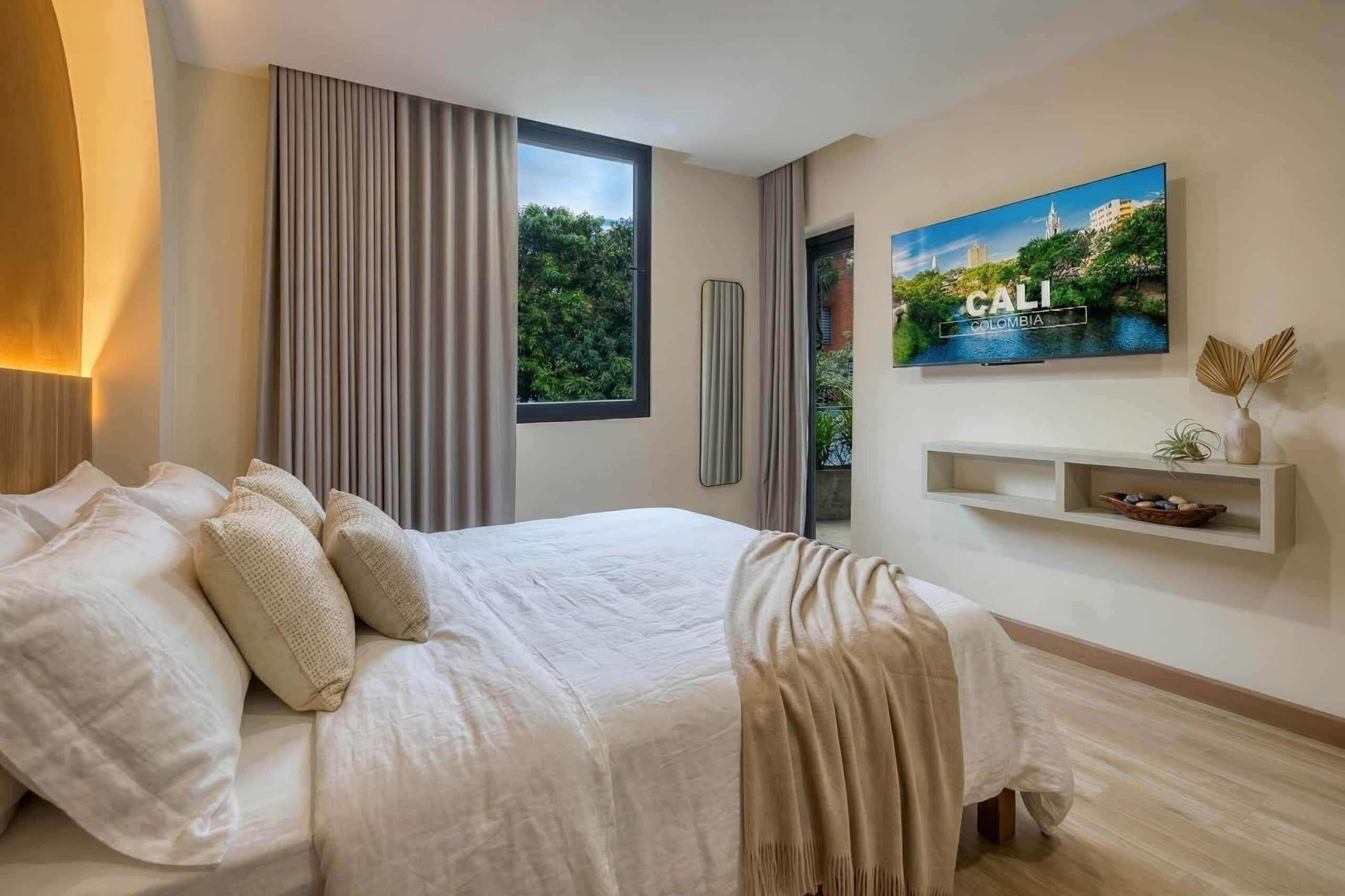 A modern bedroom with a large bed, a window with curtains, a wall-mounted TV, and a shelf with decorative items.