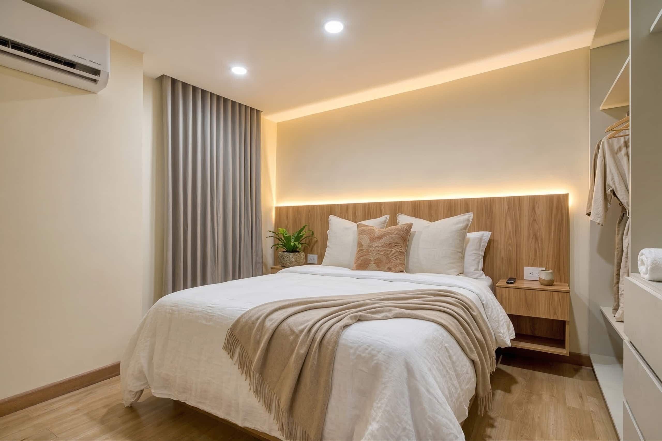 A modern bedroom with a wooden headboard, a bedside table with a lamp, and a large bed with a beige blanket.