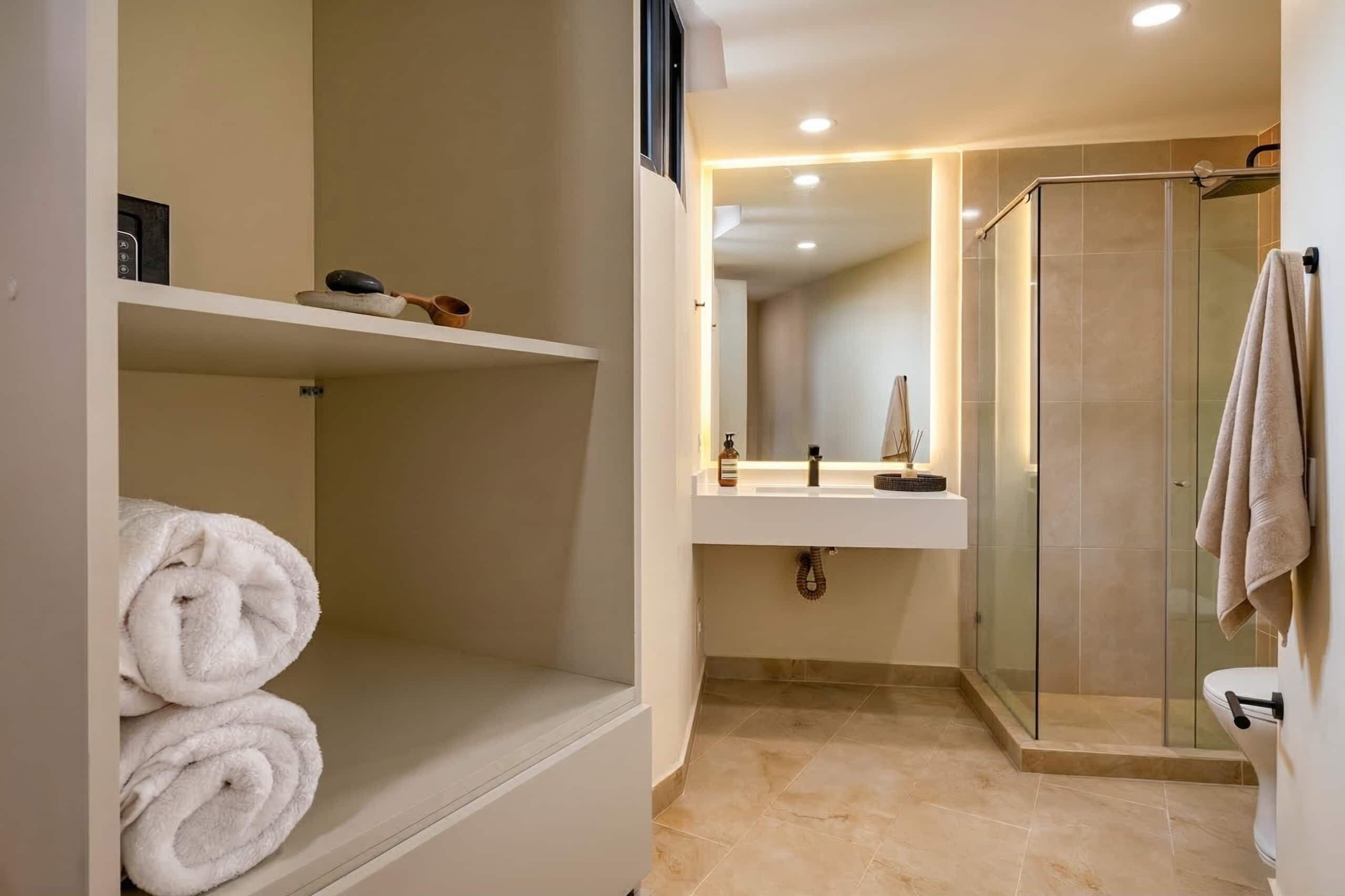A modern bathroom with a glass-enclosed shower, a sink with a mirror, and a shelf with rolled towels.