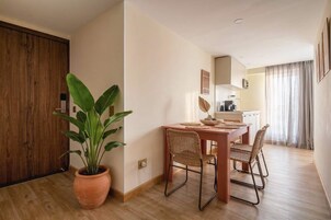 A dining area with a wooden table, wicker chairs, a potted plant, and a kitchen in the background.