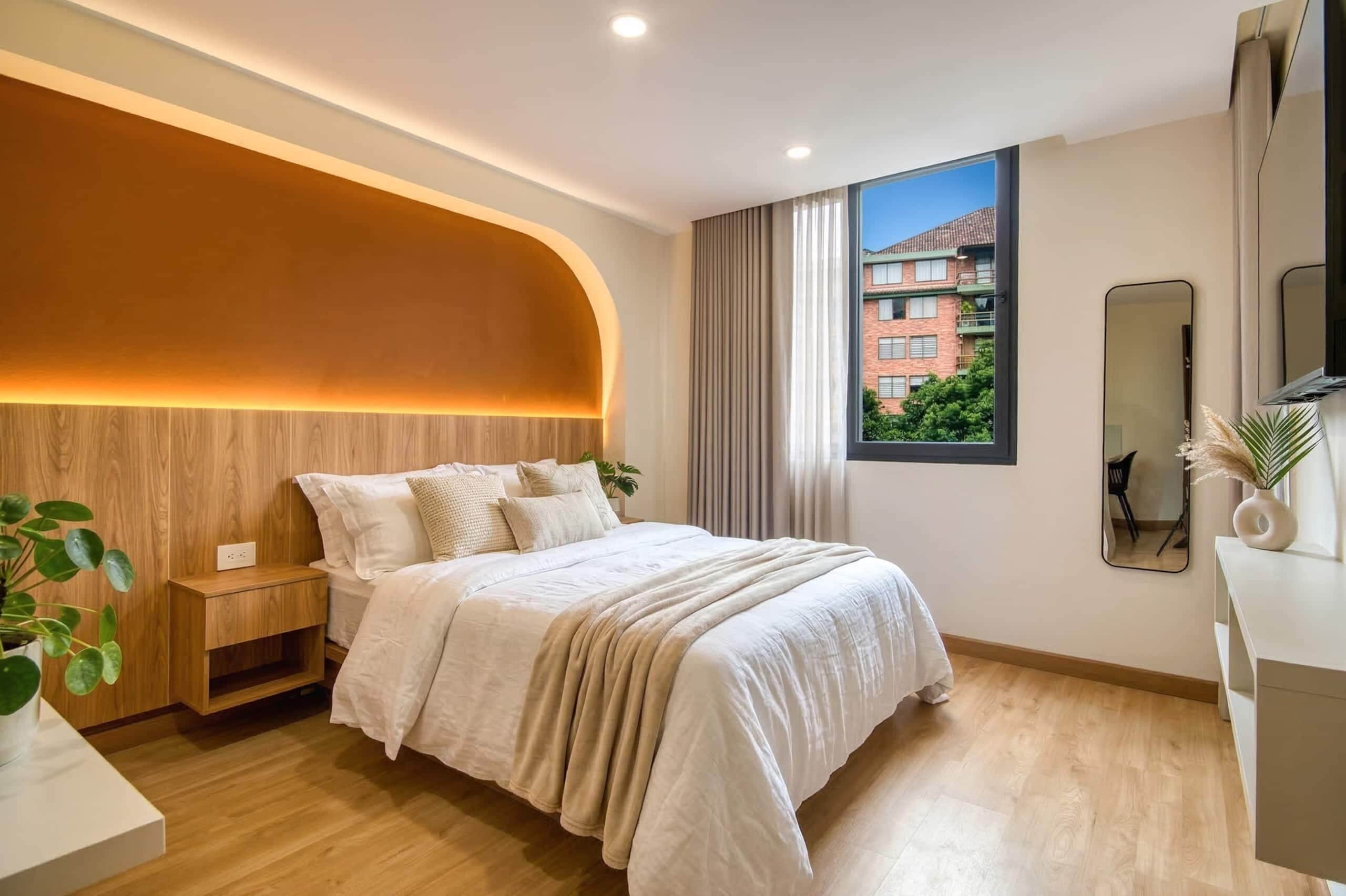 A modern bedroom with a curved headboard, a large window with a view of buildings, a bedside table with a plant, and a mirror on the wall.