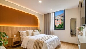 A modern bedroom with a curved headboard, a large window with a view of buildings, a bedside table with a plant, and a mirror on the wall. - Passiflora luxury suite (Cali)