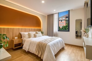 A modern bedroom with a curved headboard, a large window with a view of buildings, a bedside table with a plant, and a mirror on the wall. - Passiflora luxury suite (Cali)