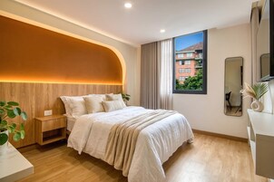 Un dormitorio moderno con una cabecera curvada, un ventanal con vistas a edificios, una mesita de noche con una planta y un espejo en la pared.