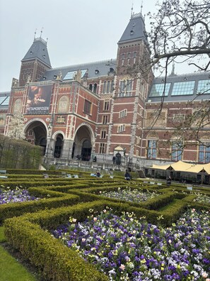 Ein historisches Gebäude mit großem Bogenportal, einem Uhrturm und einem Garten mit geschnittenen Hecken und Blumen.