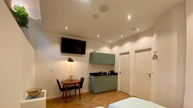 Una habitación compacta con una cama, una pequeña mesa de comedor con sillas, un televisor montado en la pared y una kitchenette con armarios.