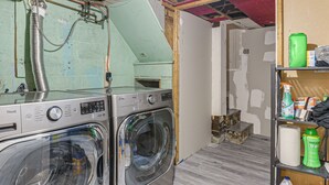 Un área de lavandería con dos lavadoras LG, un estante con productos de limpieza y una habitación parcialmente terminada con ladrillo visto y vigas de madera.