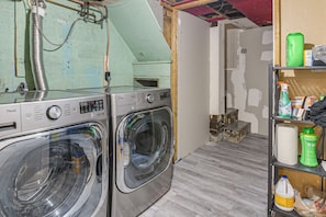 A laundry area with two LG washing machines, a shelf with cleaning supplies, and a partially finished room with exposed brick and wooden beams.