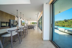Eine moderne Küche mit Barhocker, ein Essbereich mit Tischen und ein großes Fenster mit Blick auf einen Pool.