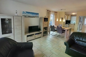 Ein Wohnzimmer mit Fernseher, eine Essecke mit Stühlen, eine Küche und ein schwarzes Ledersofa.