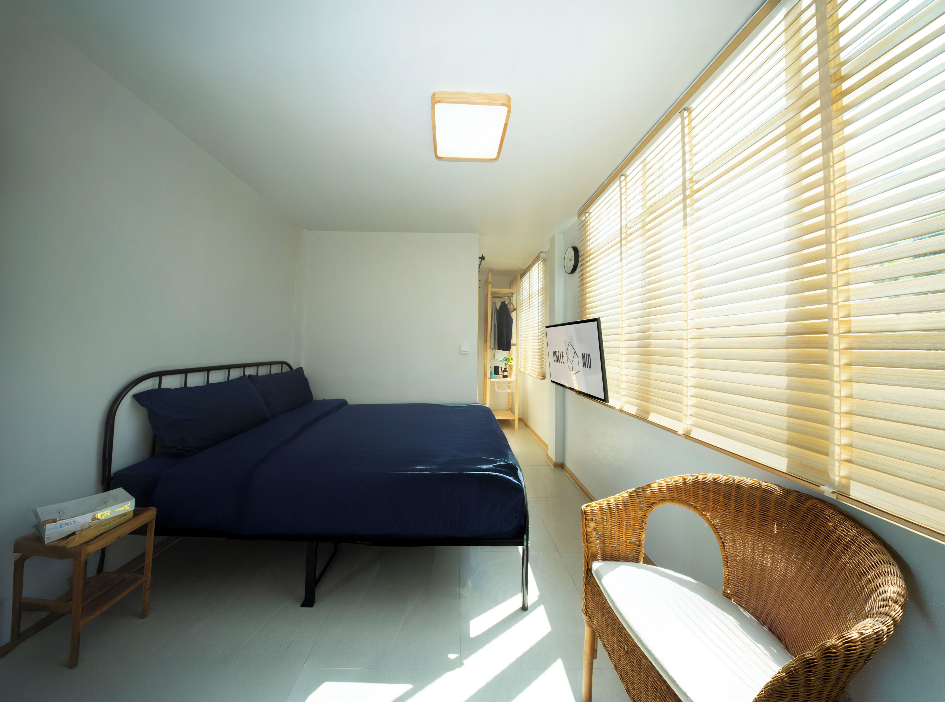 Une chambre à coucher comprenant un lit, une chaise, une table de chevet, un téléviseur fixé au mur et une fenêtre avec des stores.
