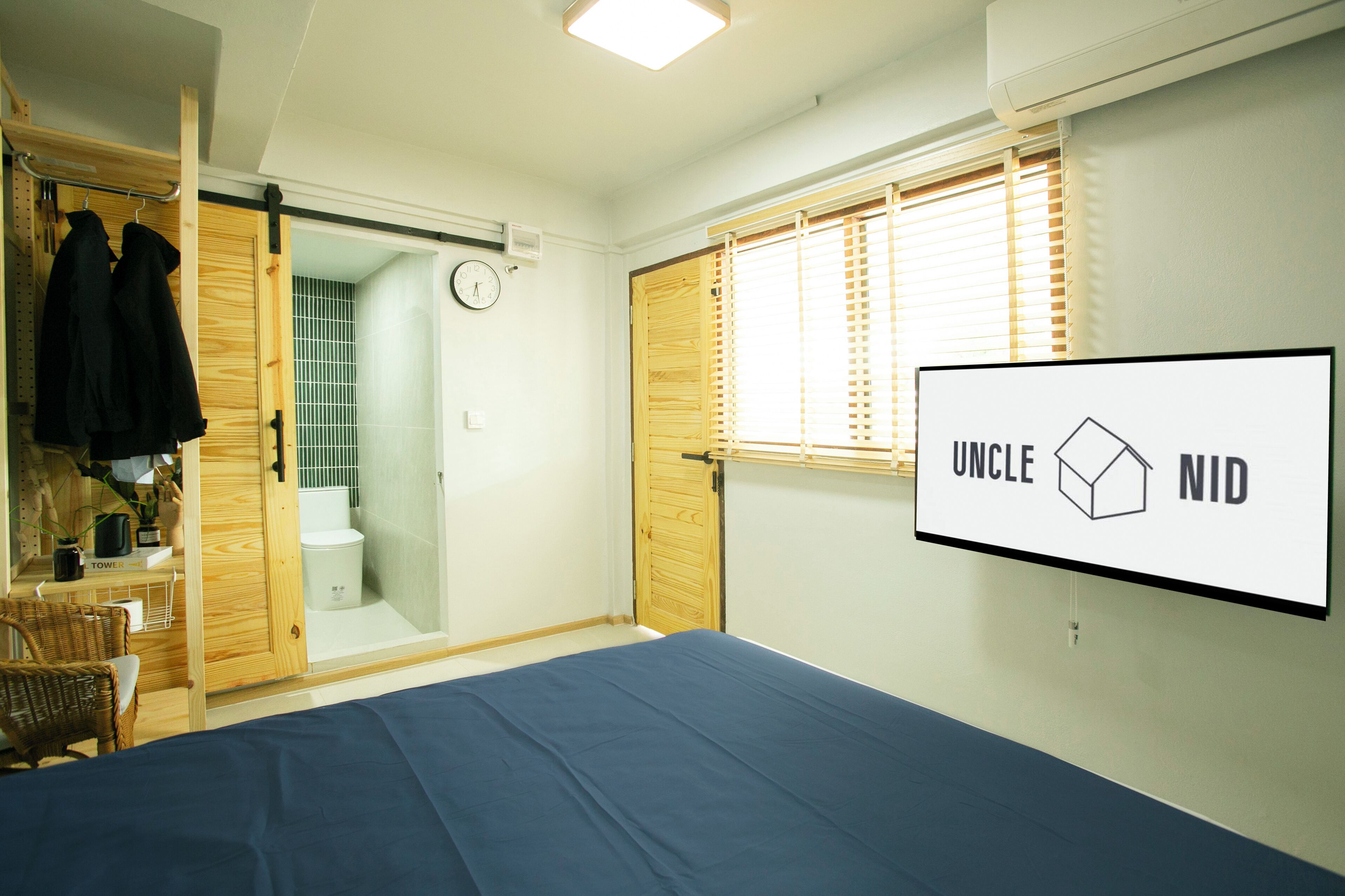 A bedroom with a bed, a wooden door, a window with blinds, a wall-mounted TV, and a clock.