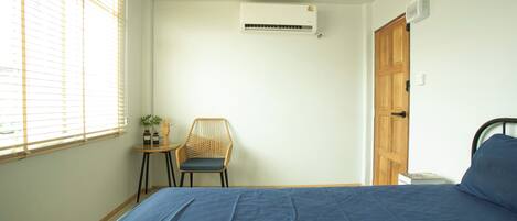 A bedroom with a bed, a chair, a small table with a plant, an air conditioning unit, and a wooden door.
