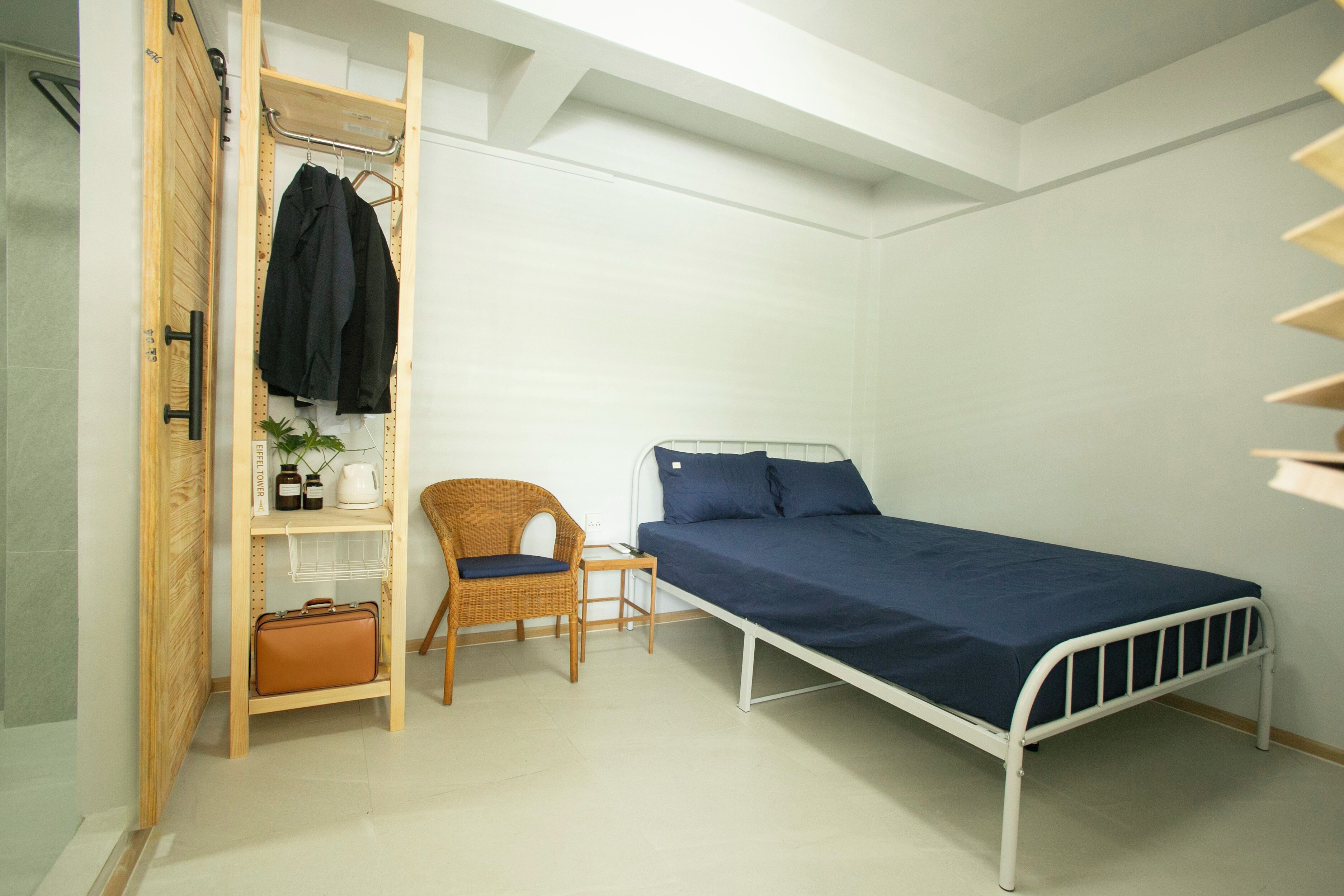 A single-bed room with a pull-out wardrobe, a wicker chair, a small table, and a suitcase on a shelf.
