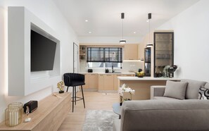 Ein modernes Wohnzimmer mit Fernseher, einer Couch, einem Couchtisch und einer Küchenzeile mit Fenster, Spüle und Schränken.