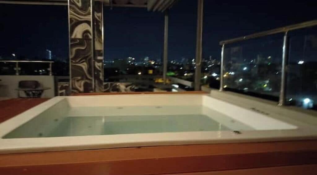 A rooftop pool with a wooden deck, surrounded by glass railings and a cityscape view.
