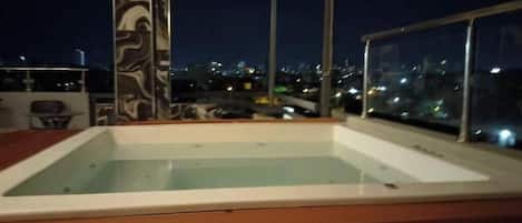 Piscina no terraço com deck de madeira, cercada por grades de vidro e vista para a cidade.