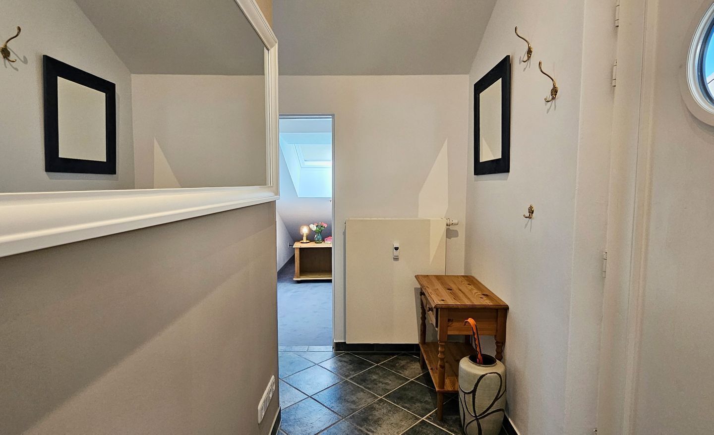 Un pasillo estrecho con paredes blancas, un banco de madera, una pequeña mesa con un florero y una puerta que da a otra habitación.