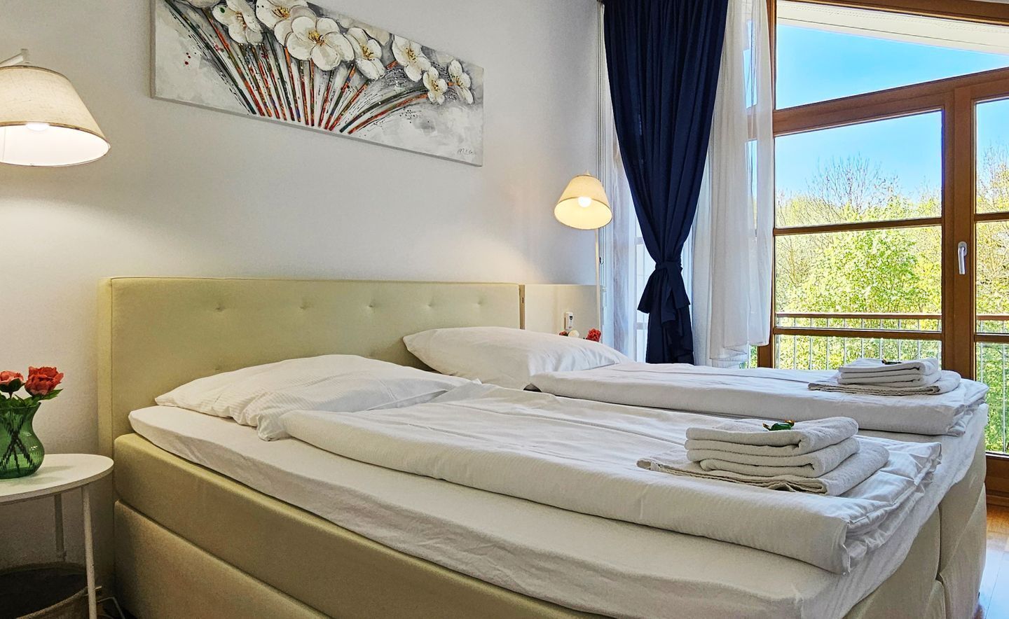Quarto de hotel com duas camas, um quadro na parede, uma janela com cortinas e um vaso com flores sobre uma mesa de apoio.