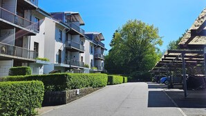 Une rue résidentielle bordée d’immeubles à plusieurs étages, avec un ciel dégagé et une zone de stationnement où sont garées des voitures.