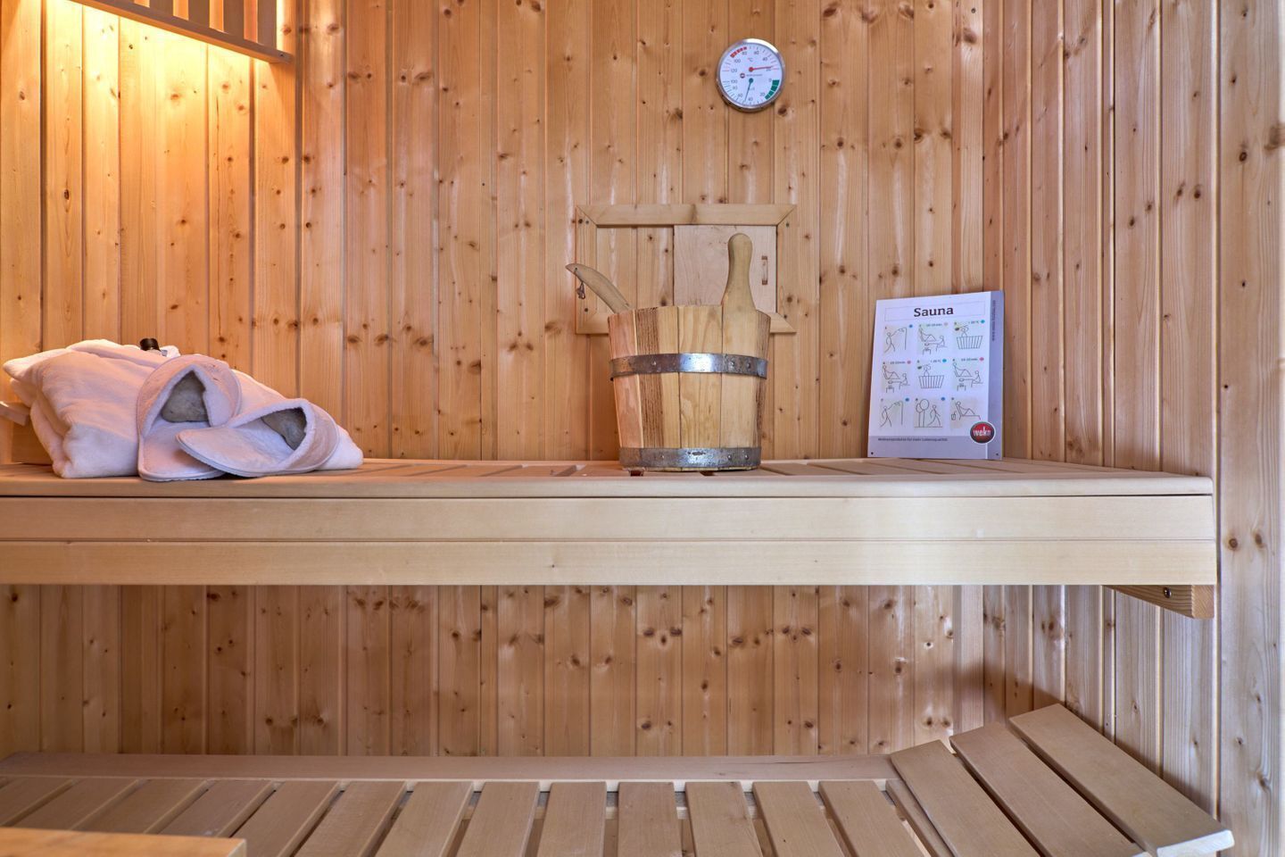 Una sauna con un banco de madera, un cubo, pantuflas y un termómetro en la pared.