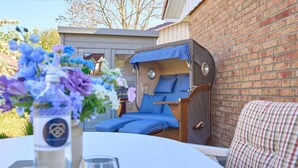 A cozy outdoor seating area with a wicker chair and a cushioned bench, a table with a vase of flowers, and a clear glass.
